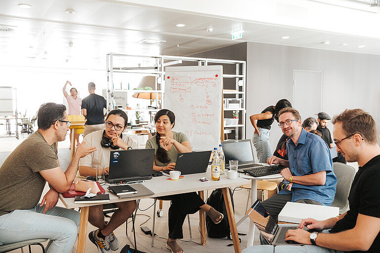 Personen mit Notebook rund um einen Tisch sitzend unterhalten sich .