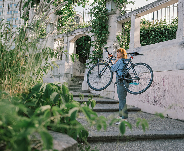 Nachhaltig Handeln: Förderungen, Beratungen, Workshops für Unternehmen. Eine Frau im Park trägt ein Fahrrad Stufen rauf