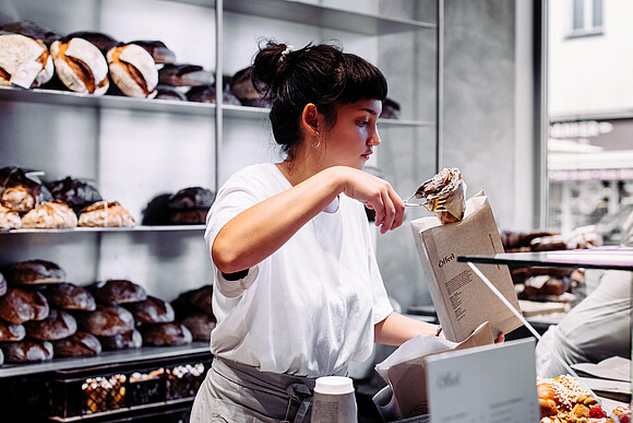 Graetzel beleben: Förderungen, Beratungen, Netzwerke für Unternehmen/Nahversorger*innen in Wien. Eine Frau in einer Bäckerei steckt ein Stück Gebäck in ein Verkaufssackerl.