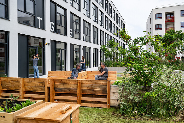 Zwei sitzende Männer im Gespräch vor dem Gewerbehof Seestadt, ein stehender junger Mann, gehende junge Frau