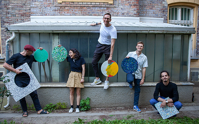 Fantoplast: Das Team von Fantoplast, eine Frau und vier Männer, stehen mit bunten runden und eckigen Paneelen in einem Innenhof.