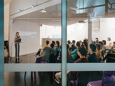 Blick durch eine Scheibe in einen Seminarraum, in dem eine Gruppe von Menschen sitzt, eine Frau mit langen blonden Haaren steht auf der Bühne und spricht.