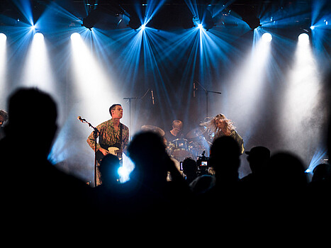 Menschen stehen vor einer Bühne, auf der drei Musiker*innen performen