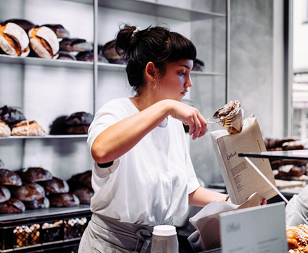 Graetzel beleben: Förderungen, Beratungen, Netzwerke für Unternehmen/Nahversorger*innen in Wien. Eine Frau in einer Bäckerei steckt ein Stück Gebäck in ein Verkaufssackerl.