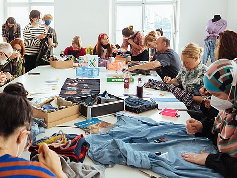 Eine Gruppe Menschen sitzt um einen Tisch auf dem verschiedene Stoffe liegen. Manche Leute nähen, andere unterhalten sich.