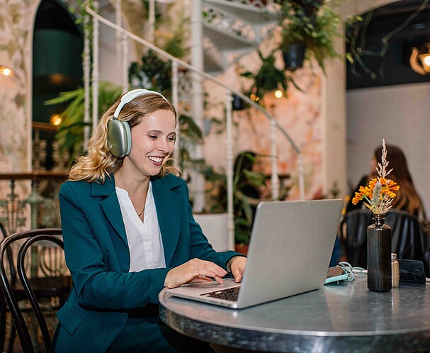 Eine Frau sitzt an einem Tisch und arbeitet an einem Laptop, am Kopf trägt sie ein Headset