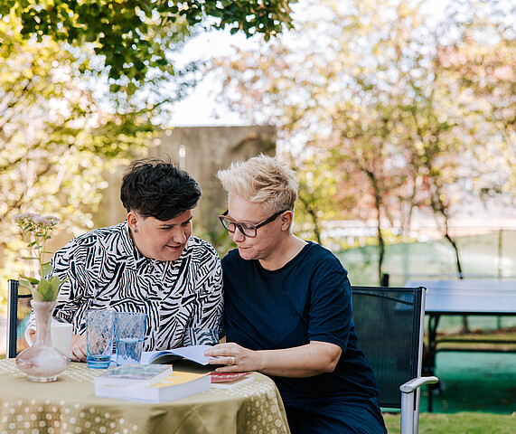 Bistra and Christina Two women sitting at a table, reading a book together.