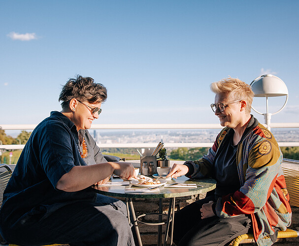 WirtschaftsagenturWien-Expat-Club Internationales Wien für Expats: Zwei Frauen sitzen auf einer Terrasse und teilen sich einen Kuchen. Im Hintergrund Skyline von Wien. Mitglied im Expat Club werden.