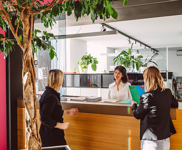 InternationalBusiness-Leben-Arbeiten-Wien Zwei Frauen werden an einem hellen Empfangsbereich von einer anderen Frau willkommen geheißen. Expat Center: Leben und Arbeiten in Wien.